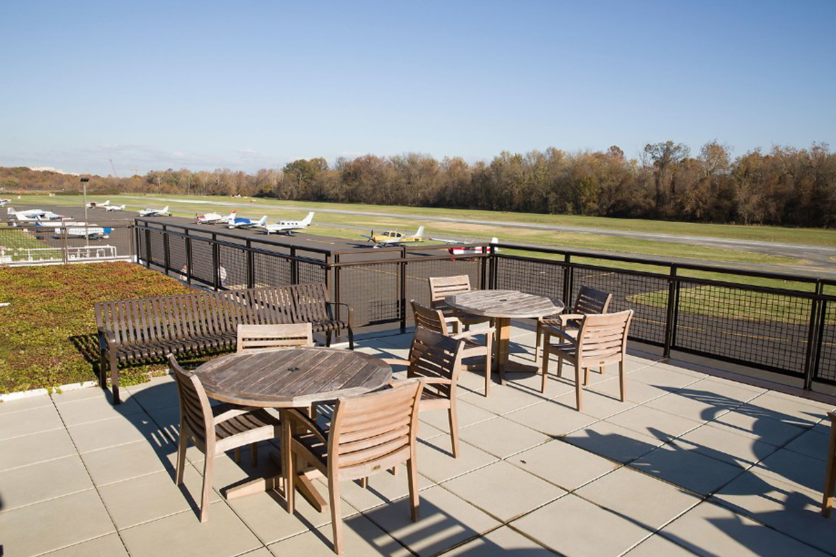 Tables and chairs on an outdoor deck overlooking a runway. Parked planes are visible in the distance.