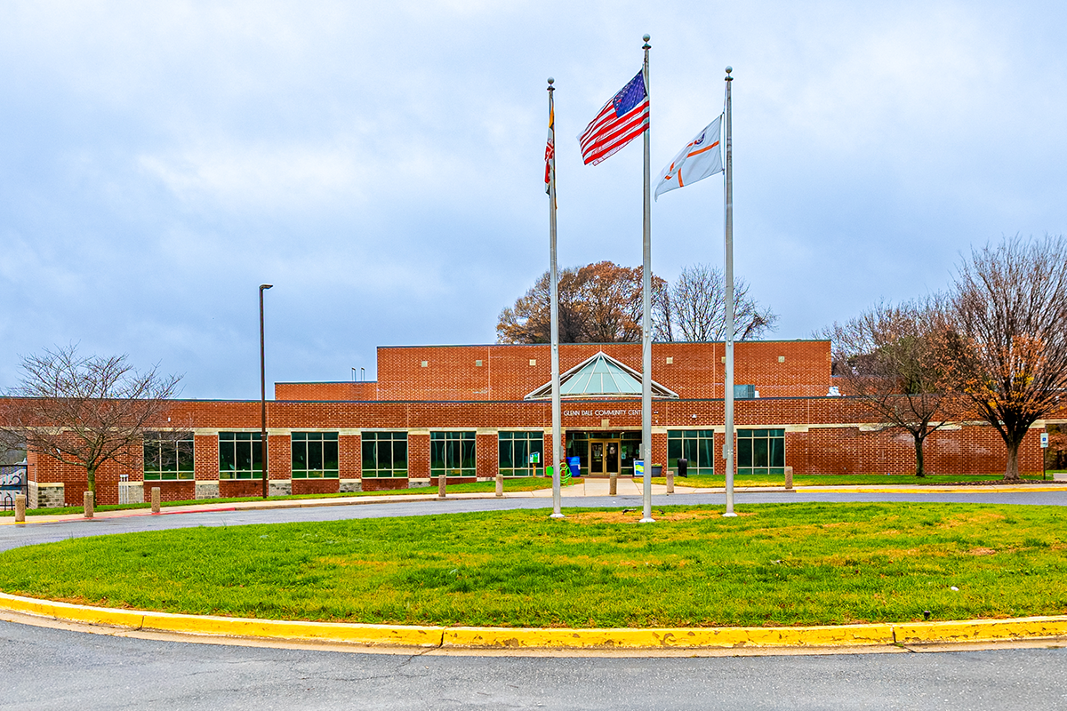 Exterior view of the circular driveway in front of a large, brick building. Inside the circular drive are three flags on flagpoles. Silver lettering over the building entrance reads Glenn Dale Community Center.