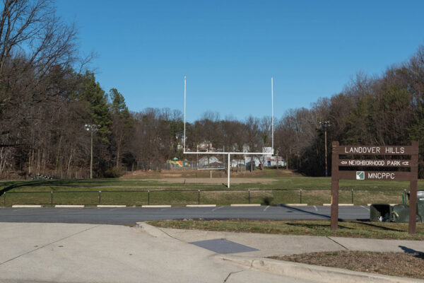 A brown wooden sign reading LANDOVER HILLS NEIGHBORHOOD PARK in the grass near a parking lot. Behind the lot is a grass field with football goalposts in it, surrounded by trees.