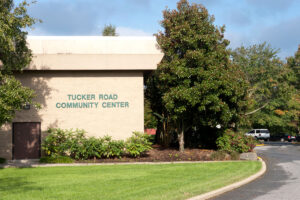 Exterior view of the corner of a beige brick building with trees around it. Green lettering on the building reads TUCKER ROAD COMMUNITY CENTER.