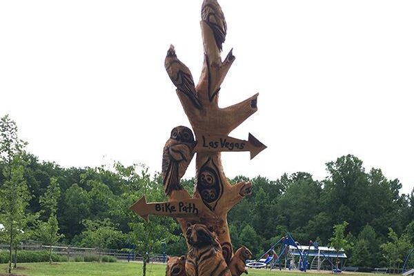 A vertical outdoor sculpture resembling forest animals perching on a tree trunk. Directional signage arrows on the sculpture are labeled Bike Path and Las Vegas. In the background is an expanse of grass, a playground, and trees.