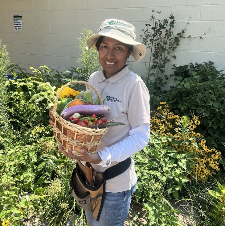 A smiling staffer stands outside in a garden holding a basket of vegetables, including eggplant, tomatoes, and bell peppers.