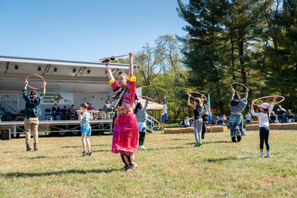 Adults and kids dance with plastic hoops in the grass in front of a portable bandstand with musicians. A sign in the bandstand reads 