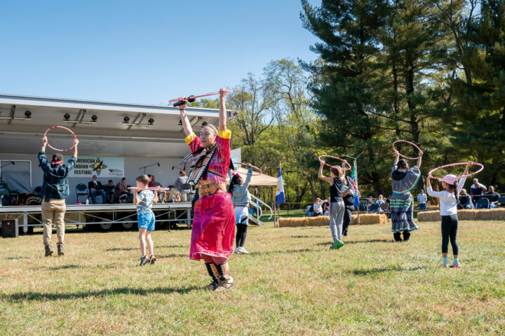 Adults and kids dance with plastic hoops in the grass in front of a portable bandstand with musicians. A sign in the bandstand reads