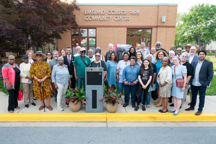 About 35 people gather together for a photo on a sidewalk in front of a brick building with lettering that reads LAKELAND COLLEGE PARK COMMUNITY CENTER. The group stands behind a podium flanked by two potted plants; posters on an easel are situated nearby.