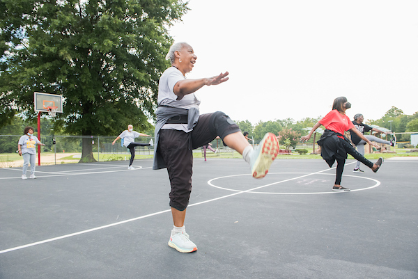 A group of senior women kick their legs high in a fitness class being held on an outdoor basketball court.