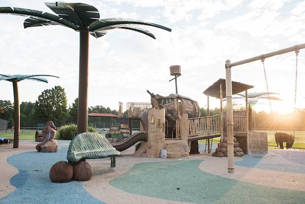 An outdoor playground with a shipwreck/desert island theme, featuring artificial palm trees, a sand castle play structure, a multi-level shipwreck play structure, swings, and more. Beyond the playground are tennis courts and baseball fields.