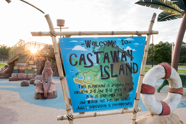At an outdoor playground, a large sign on fake bamboo poles reads "WELCOME TO CASTAWAY ISLAND. This playground is designed for children 2-5 and 5-12 years of age." Behind the sign, elements of a shipwreck-themed playground are visible, including a play structure topped with ship's mast and crow's nest, coconut stepping stones, and an artificial palm tree.