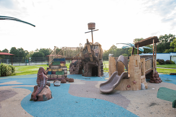 An outdoor playground with a shipwreck/desert island theme, featuring multiple slides, climbing structures, artificial palm trees, and an alligator sculpture. The central play structure resembles a wrecked ship. Back behind the playground are tennis courts, shade structures, and a restroom/comfort station building.
