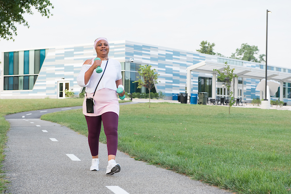 A smiling woman wearing pink work-out clothes and holding small blue weights in both hands walks along a paved trail with a white dotted line down the middle. The trail is leading away from a large, modern, blue, gray, and white-tiled building.