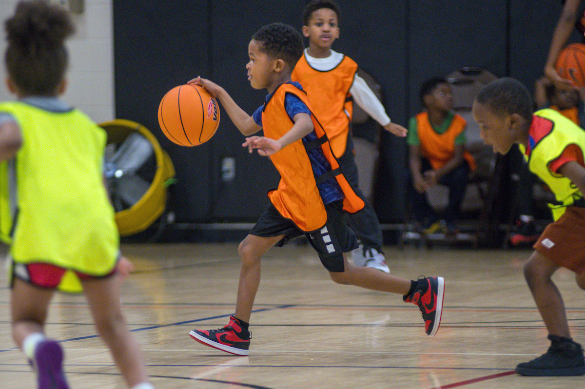 Inside a gymnasium, a boy wearing black and red Air Jordans and an orange smock over his clothes runs while dribbling a basketball. He is pursued by a member of the opposing team in a yellow smock.
