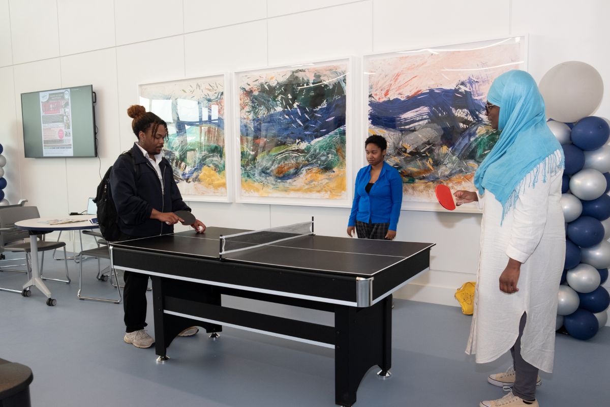 Inside a lounge, a man and woman play ping pong on a small, black table, while another woman watches. There are a balloon sculptures in the corners of the room.