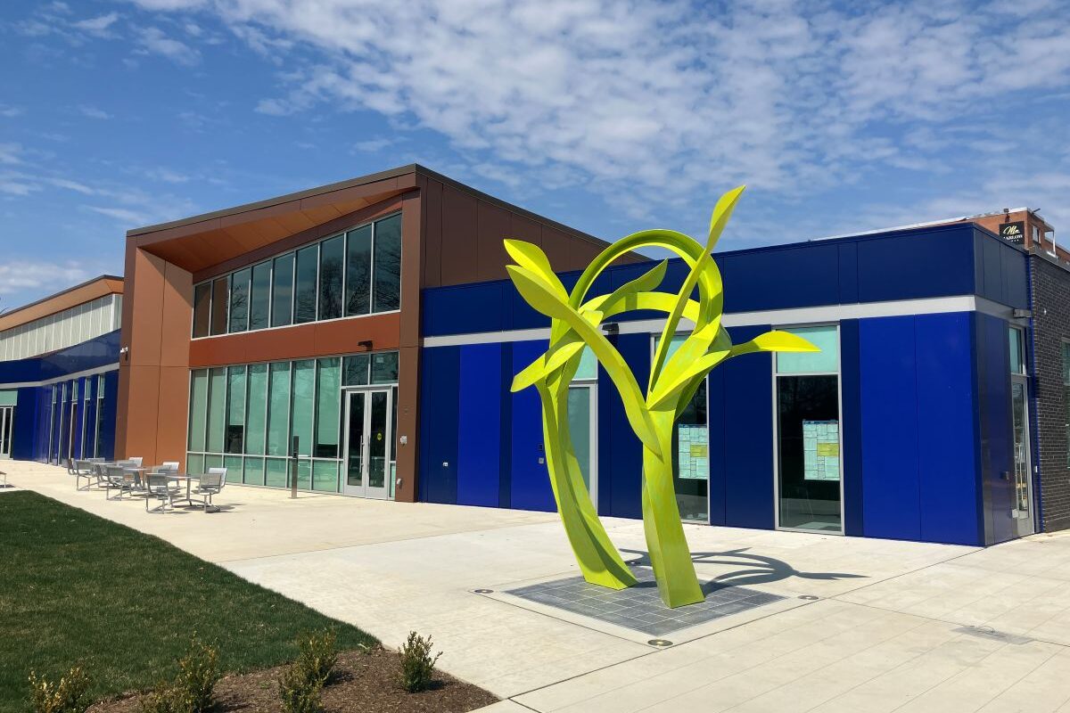 Exterior view of a large, modern, glass, brick, and metal building. Some sections of the building are royal blue. Out in front is a large, neon green sculpture as tall as the building, that resembles plant stems and leaves.