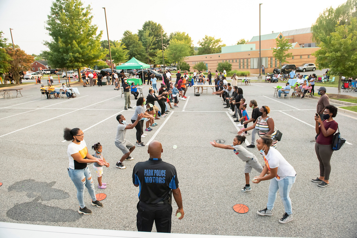 In a parking lot outside a large, brick building, two rows of people stand apart, tossing water balloons back and forth, at a community event.