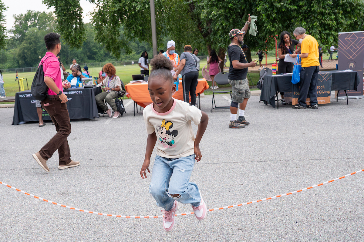 A girl wearing a Minnie Mouse t-shirt jumps rope in a parking lot. Behind her, where the parking lot meets a grassy field, are vendors and information tables.