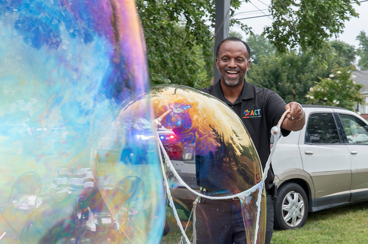 Outside in a grassy area, a smiling man wearing a black polo shirt creates giant rainbow soap bubbles using a loop of rope attached to two handles.