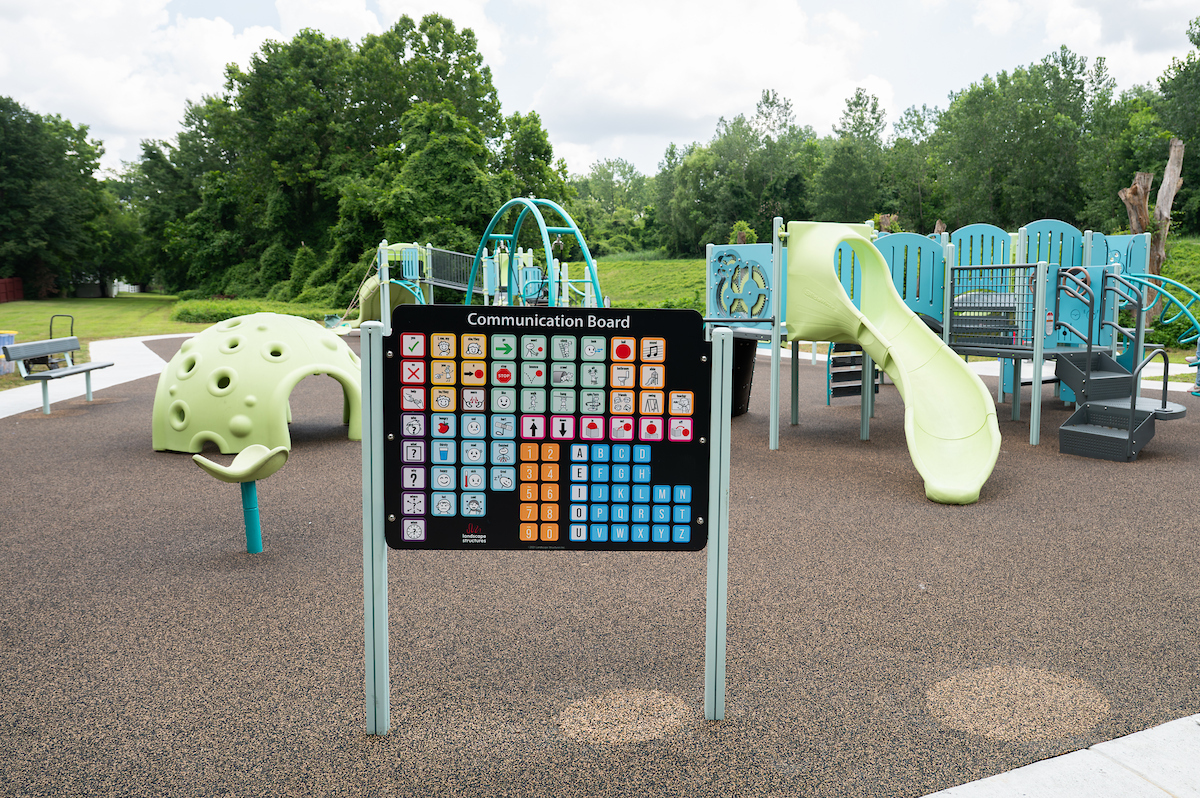 At an outdoor playground, a Communication Board is installed near the edge, containing various icons and images as well as the alphabet and numbers. The playground features slides, platforms, monkey bars, spinning elements, swings, rope webs, and more.