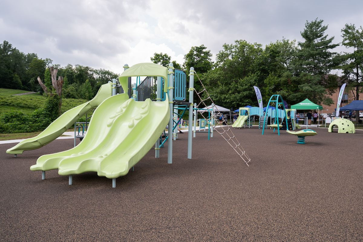 An outdoor playground with multiple play structures, including swings, slides, rope webs, monkey bars, platforms, rocking elements, and spinning elements. Beyond the playground are grass and trees.