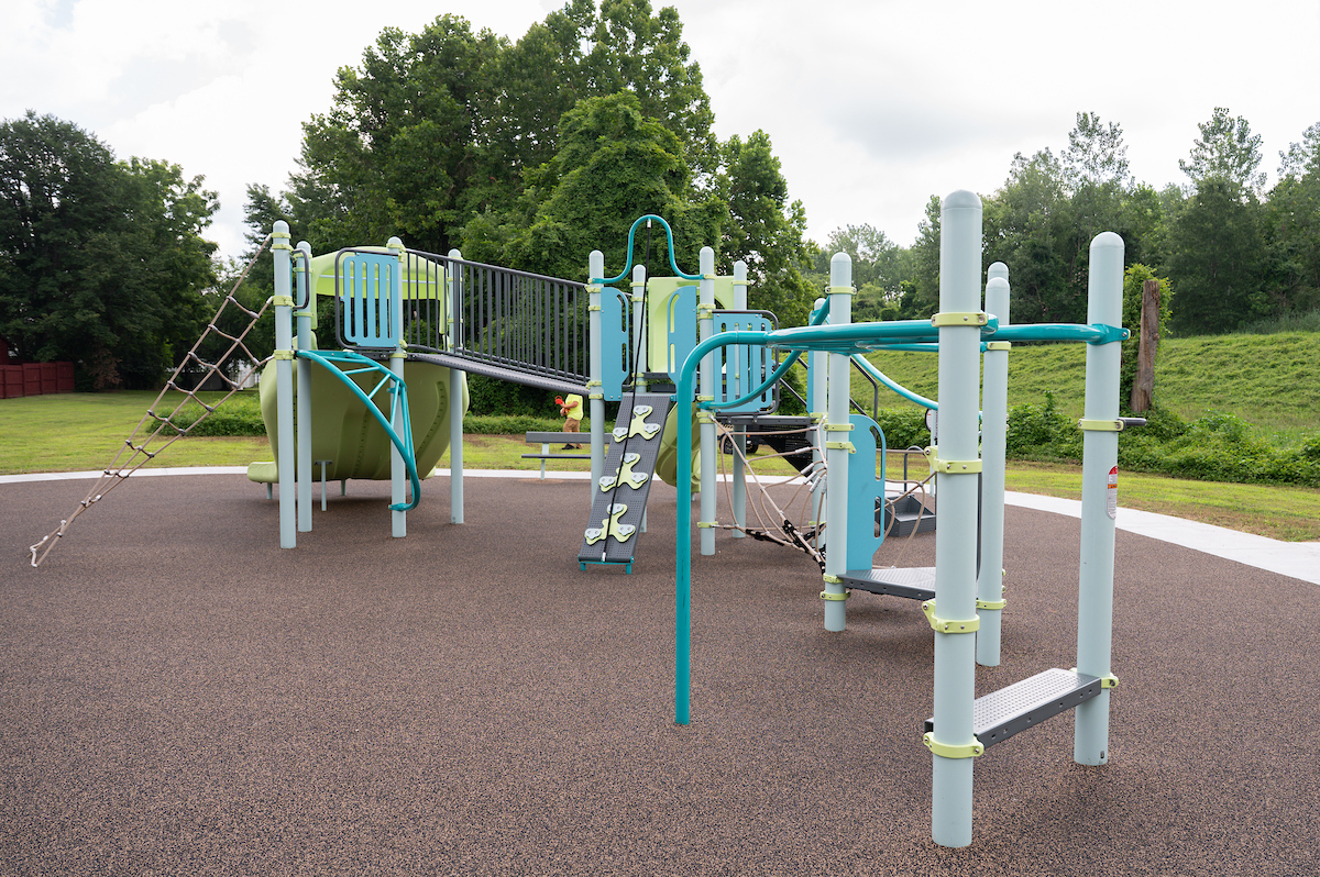 An outdoor playground with a large play structure featuring rope webs, monkey bars, slides, a climbing wall, platforms, a bridge, stairs, and more. The playground is green and blue with gray vertical posts.