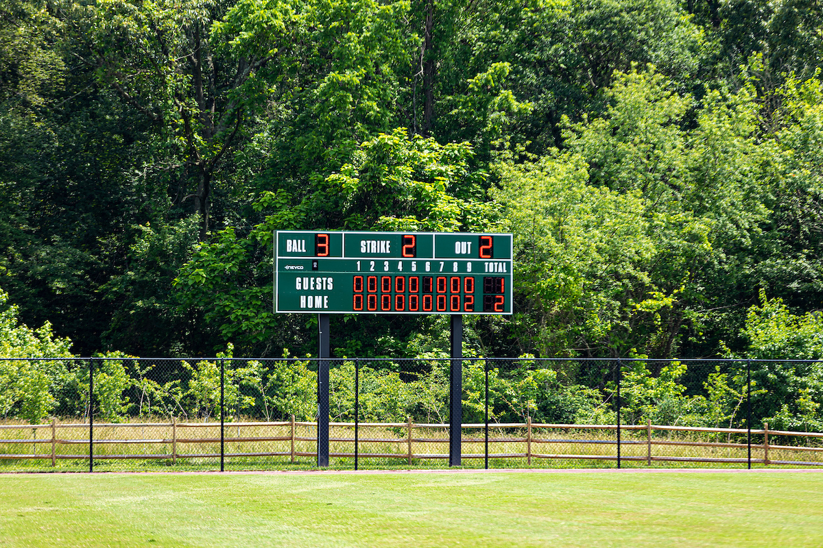 A scoreboard installed at the fenced outfield edge of a baseball diamond. Beyond the fence are tall tees.