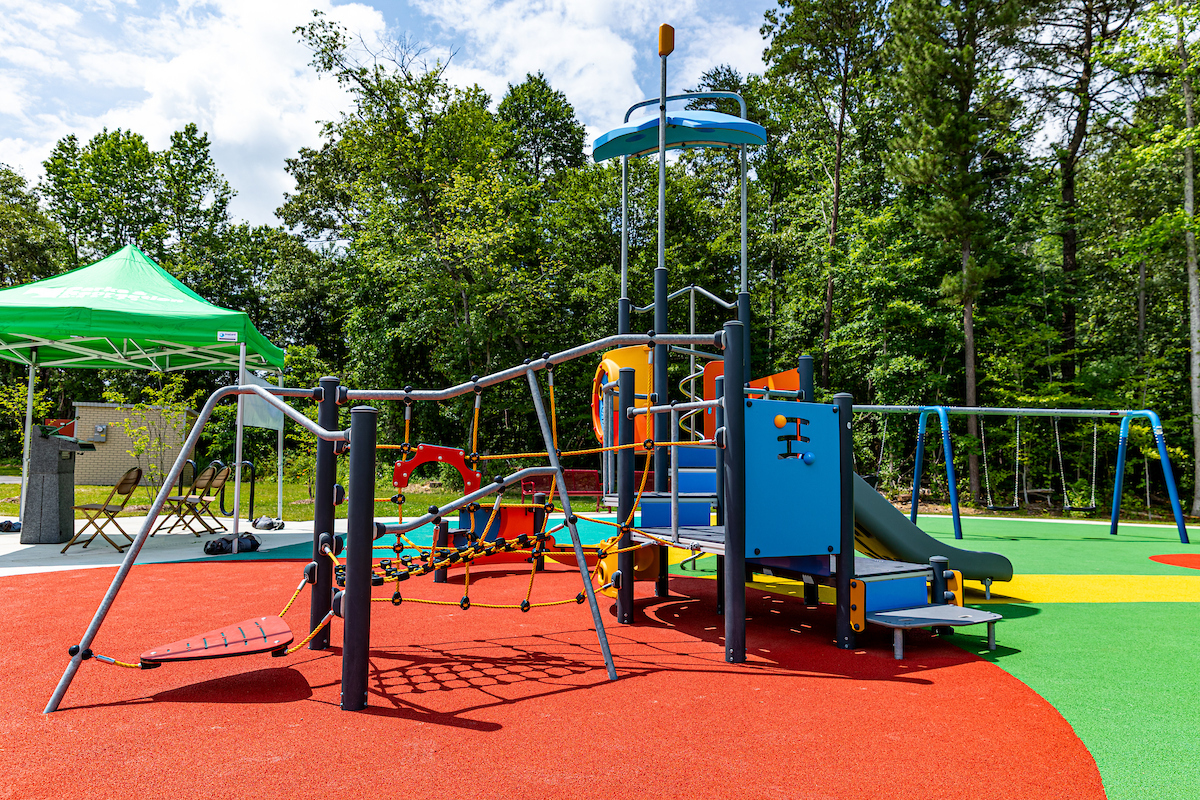 A colorful outdoor playground featuring swings, slides, rope webs, platforms, stairs, interactive toys, and more. Beyond the edge of the playground are tall trees.
