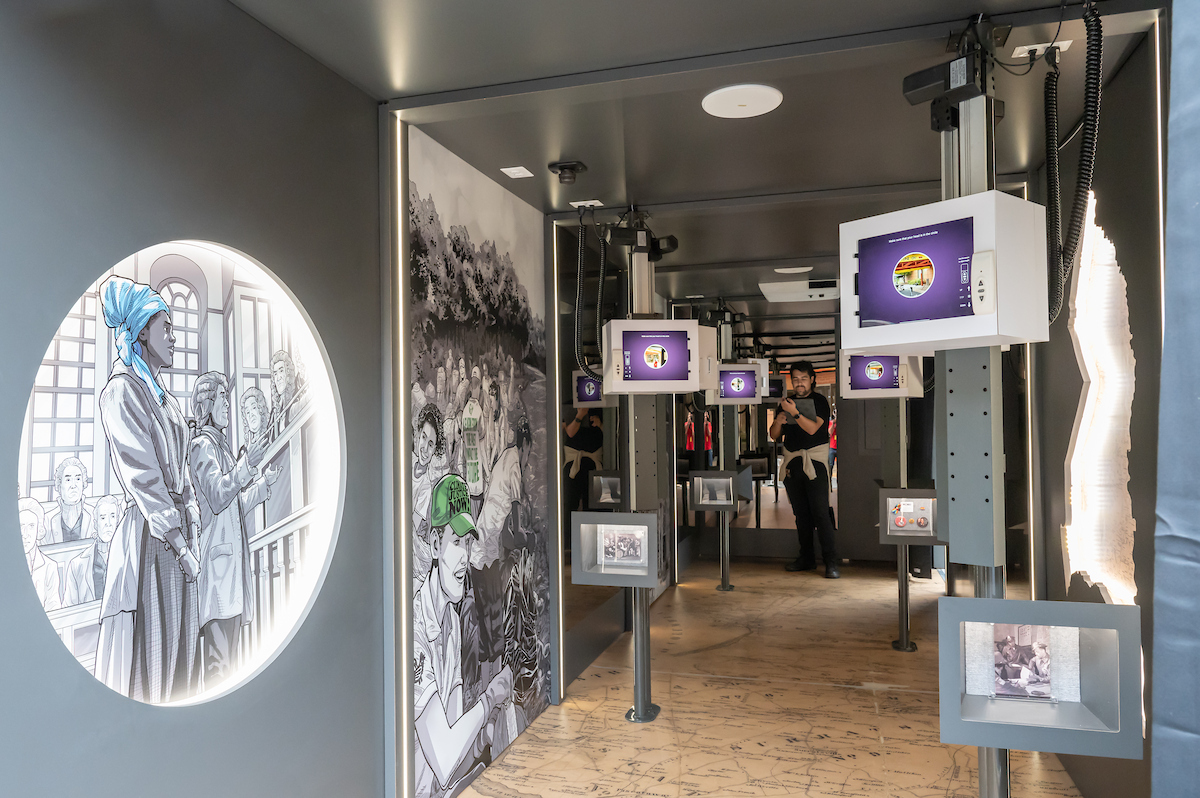 A view into a long, mobile museum containing interactive video monitors, small shadow-box exhibits, and illustrated panels along the walls. The floor is a map of Prince George's County.