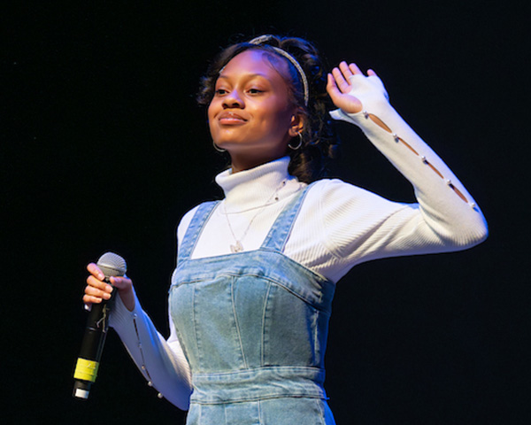 A young woman wearing denim overalls and a white turtleneck holds a microphone in her right hand, smiling. Her left arm is raised as if waving.
