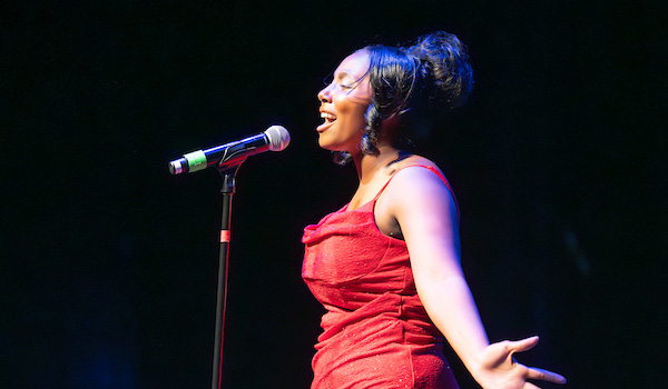 Side view of a young woman in a red formal dress singing into a microphone on a stand in front of her. Her arms are extended out to the sides in a dramatic gesture.