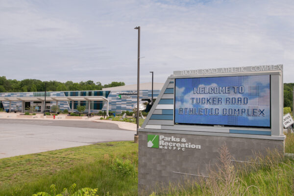 On a grassy slope is a large LED sign on a cinderblock base. Letters along the top frame of the sign read TUCKER ROAD ATHLETIC COMPLEX. The LED screen displays a photo of clouds in a blue sky, and the words WELCOME TO TUCKER ROAD ATHLETIC COMPLEX. In the background is a large, modern building with a blue, gray, and white tiled pattern that is repeated on the edge of the sign.