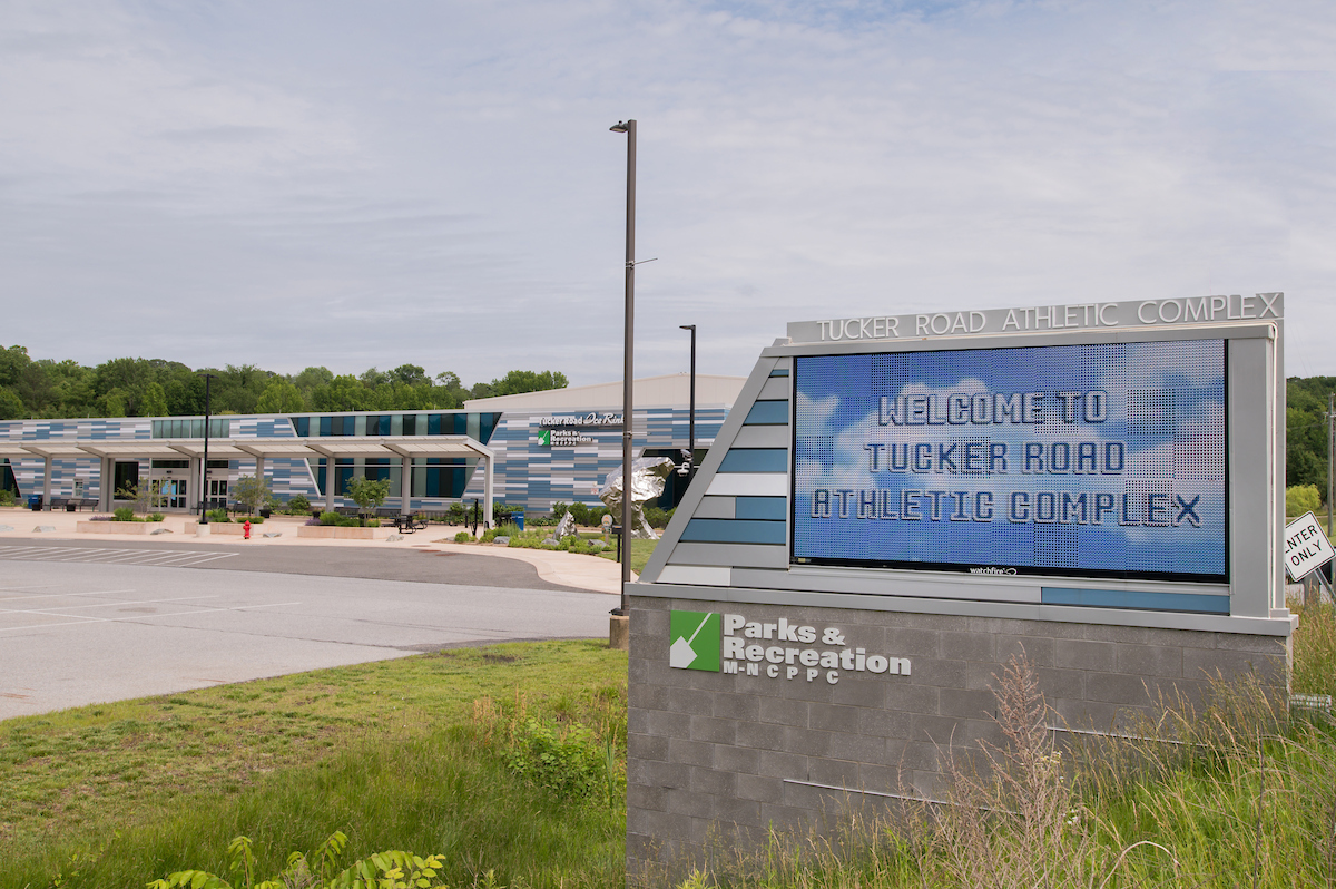 On a grassy slope is a large LED sign on a cinderblock base. Letters along the top frame of the sign read TUCKER ROAD ATHLETIC COMPLEX. The LED screen displays a photo of clouds in a blue sky, and the words WELCOME TO TUCKER ROAD ATHLETIC COMPLEX. In the background is a large, modern building with a blue, gray, and white tiled pattern that is repeated on the edge of the sign.