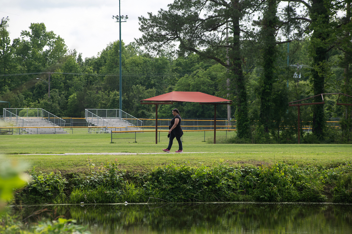 A woman in black walks along a paved trail that runs through the grass. On one side of the trail is a pond, and off the other side is an athletic field with two large sets of bleachers.