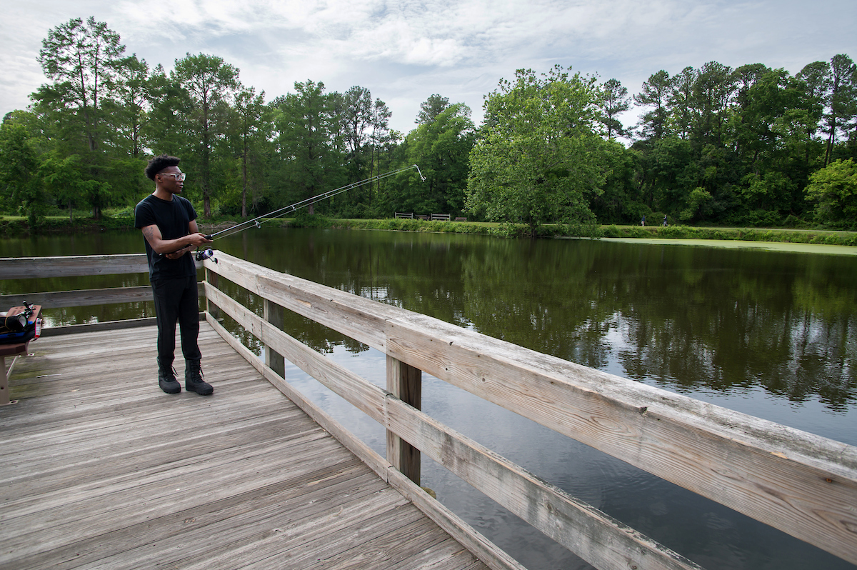 A young man wearing all black and holding a fishing pole stands on a wooden deck that overlooks a pond. He appears to be about to cast his line into the water. At the far bank of the pond are trees, grass, and wooden benches.