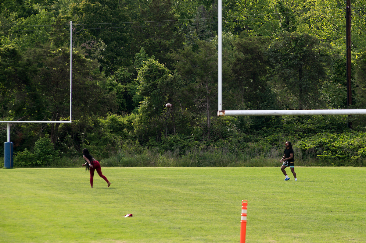 On a grass-surface football field with white goal-posts at both ends, a young woman in red leggings throws a football to a man wearing black.