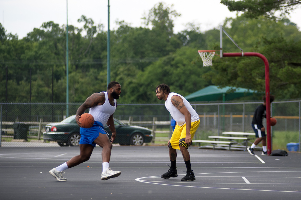 Two men in shorts and white tank tops play basketball on an outdoor, asphalt court surrounded by a chain-link fence. Outside the fence is a parking lot, a shade structure, a grassy field, and trees.