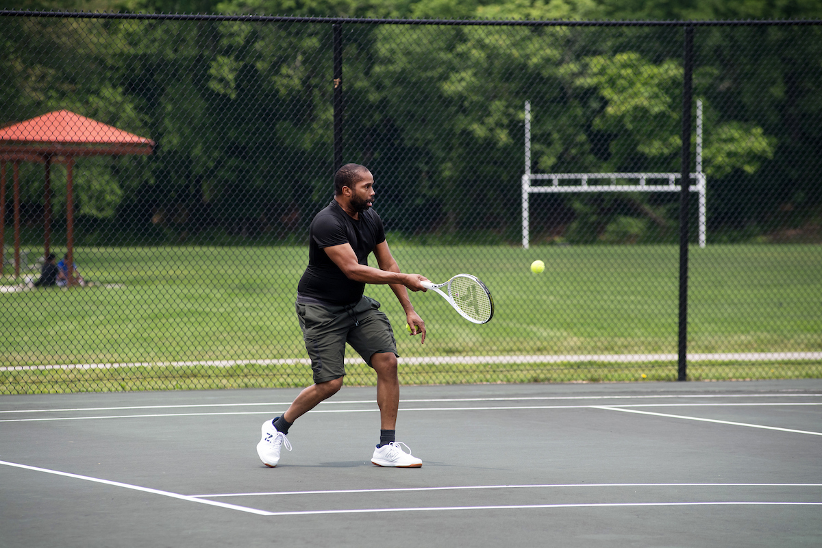 A man plays tennis on an outdoor court surrounded by a chain-link fence. Outside the fence is a green field with football goalposts at one end, and a nearby shade structure.