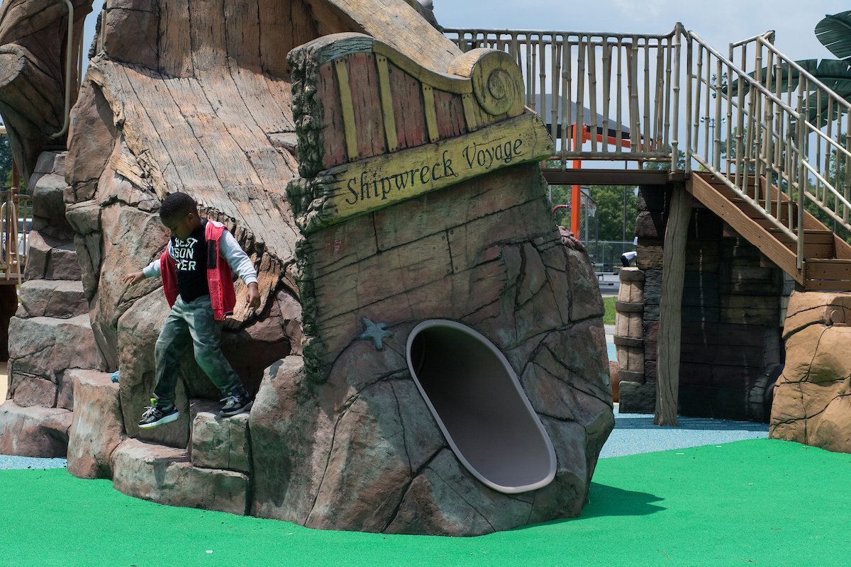 A small boy jumps off a large playground structure that resembles a shipwreck, with multiple levels, platforms, slides, and climbing features. A sign over a tunnel in the side of the playground structure reads "Shipwreck Voyage."