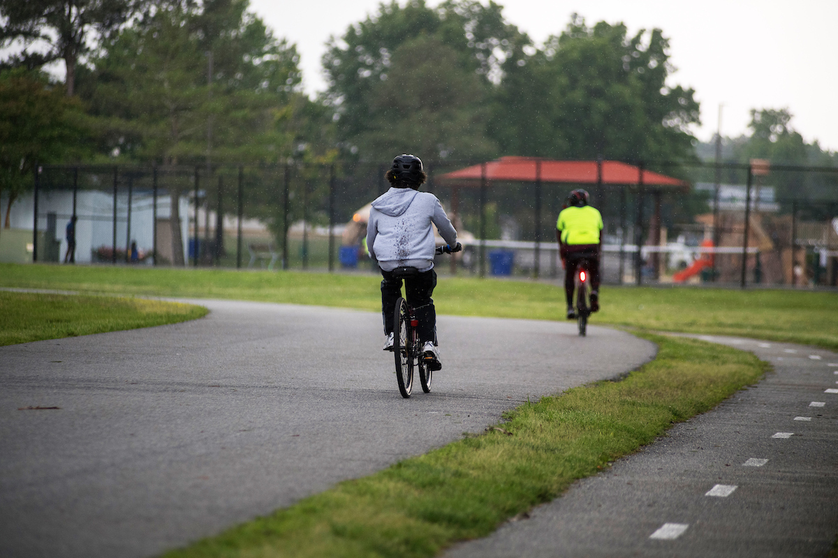 Two people ride bicycles on a paved road that runs alongside a paved trail with a dotted white line down the middle. In the blurry distance is a fitness area surrounded by a chain-link fence, and a large playground.