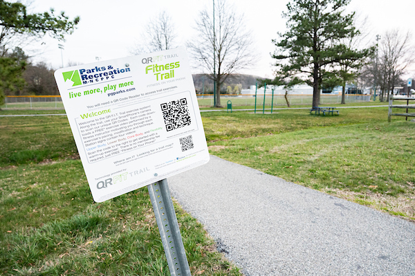 Next to a paved trail, a metal sign in the grass reads FITNESS TRAIL with a QR code on it. In the background are bleachers, picnic tables under trees, athletic fields, and a pull-up bar station along the fitness trail.