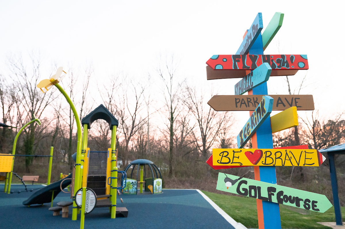 Near a green and blue outdoor playground is a wooden signpost with eleven hand-painted directional signs nailed around it at different heights. The arrow signs include locations like GOLF COURSE, ICE RINK, and PARK AVE along with motivational phrases like FLY HIGH and BE BRAVE.
