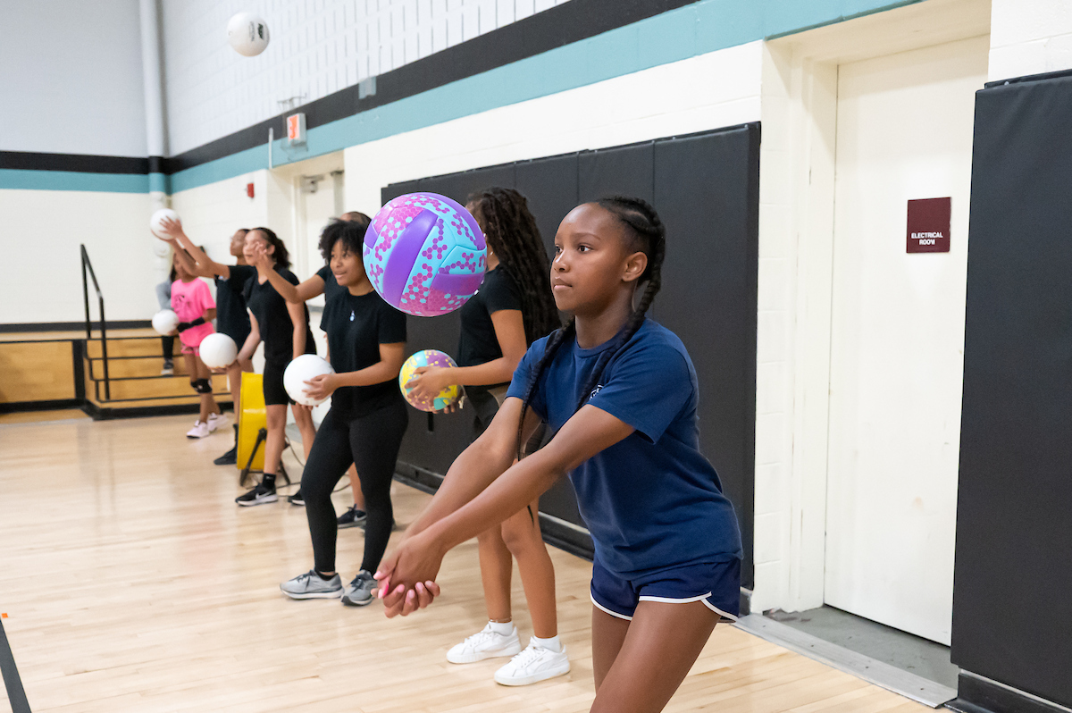 In a gymnasium, seven teen girls stand near the padded wall in a line, each with a volleyball. The girl closest to the camera has her arms out and hands laced together while practicing hitting her ball in the air, with a concentrated look on her face.