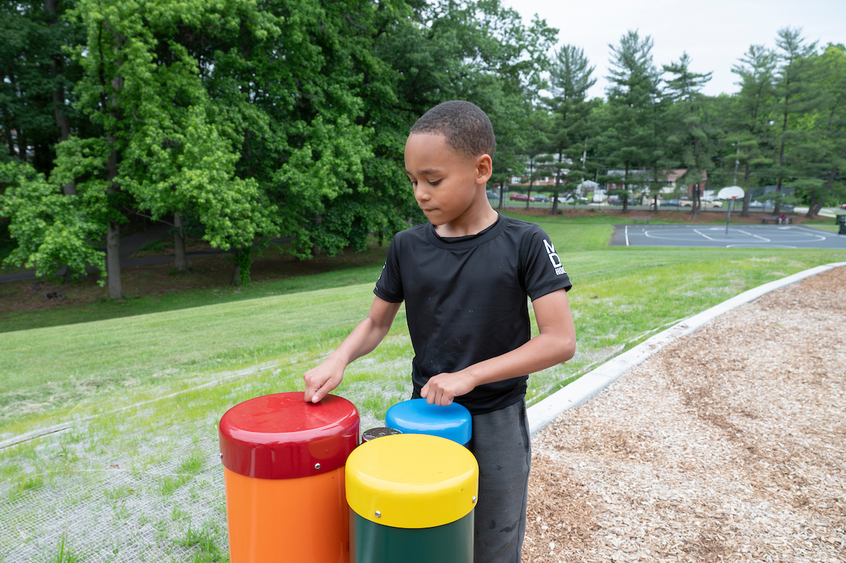 A boy in a black t-shirt drums with his knuckles on three colorful bongo-style drums at the edge of an outdoor playground. To his right is a wooded area; behind him is a basketball court.