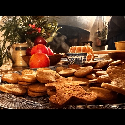 Close-up view of a plate of gingerbread cookies sitting on a table near a steaming teacup, a bowl of apples, and a ceramic, colonial-style stein. In the background is an open hearth.
