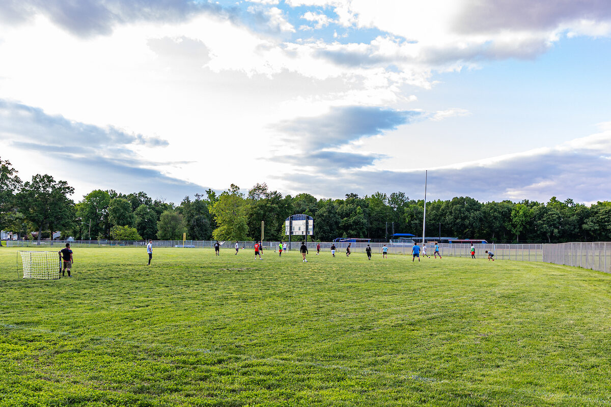 Under a blue sky with some dramatic cloud formations, people play soccer on a grass field. There are trees in the background.