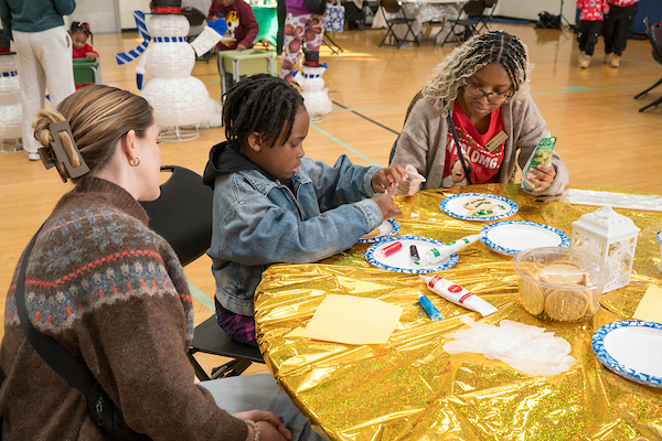 In a gymnasium, two adults sit with a child at a table with a glittery gold tablecloth. On the table are paper plates, tubes of colored frosting, and a tub of sugar cookies. In the background are more people and some snowman decorations.