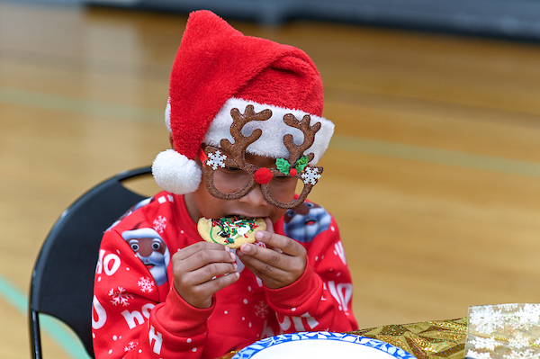 In a gymnasium, a small child wearing a red sweatshirt, a red Santa hat, and novelty Rudolph the red-nosed reindeer glasses sits at a table eating a decorated Christmas cookie.
