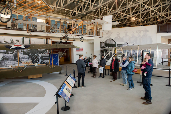 In a large, hangar-like museum gallery space with historical aircraft suspended from the ceiling and sitting on the floor, about seven people stand in a semi-circle around one man who is speaking. The nearest aircraft is a green biplane with a star painted on the top.