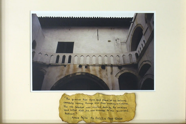 A framed photo above a scrap of paper with handwritten text. The image is of an old building with a balcony, and many archways. The text is difficult to read, but seems to say "The governor then came and stared at his balcony carefully looking through and then making his choice. The one selected was washed down by the soldiers and after that she was escorted to the governor's bedroom. - Kamia Pajibo, The Realities Abot Elaine"