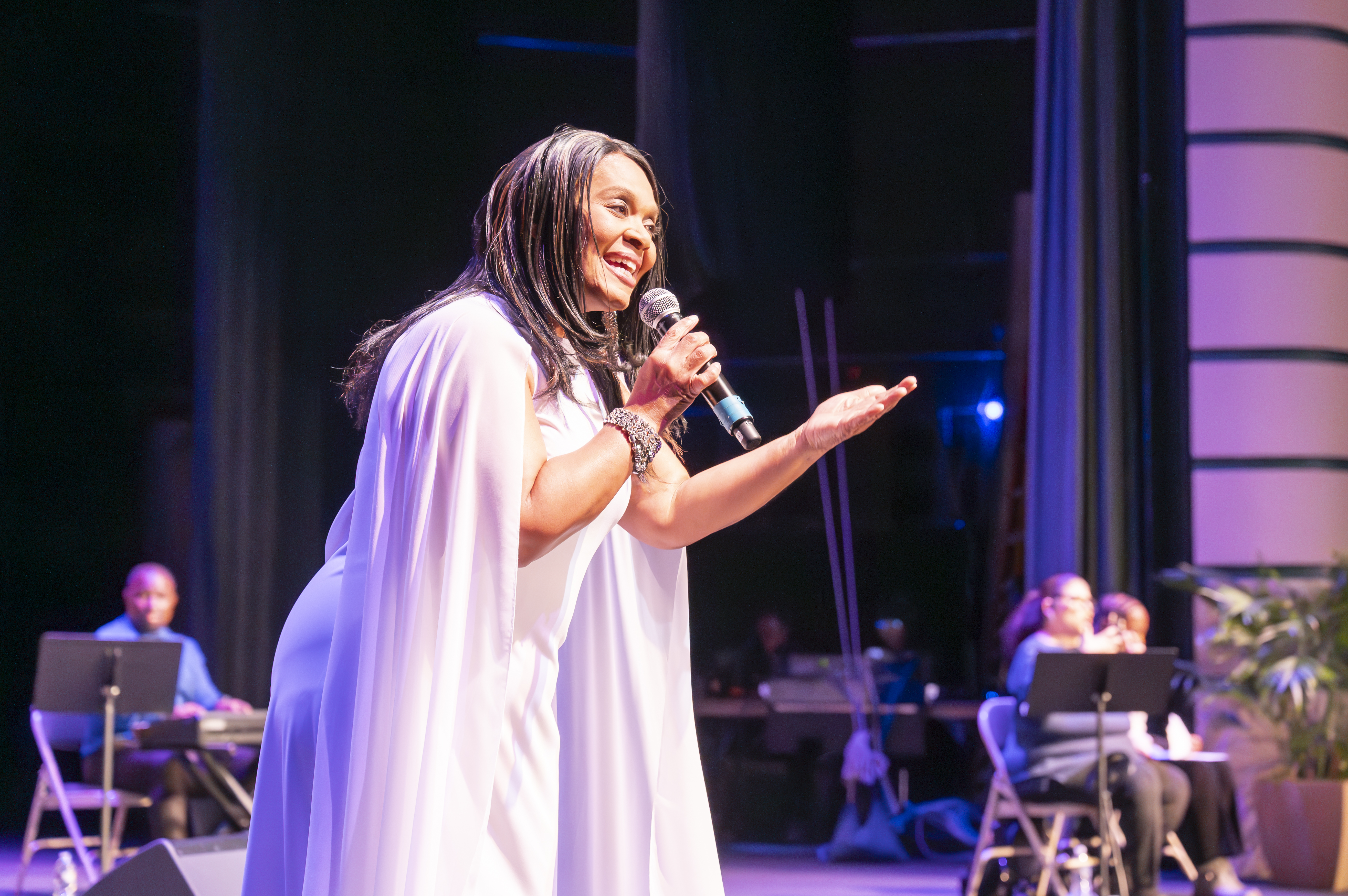 A woman in a long white dress sings into a handheld microphone on a stage. Behind her is a seated keyboard player looking at a music stand.