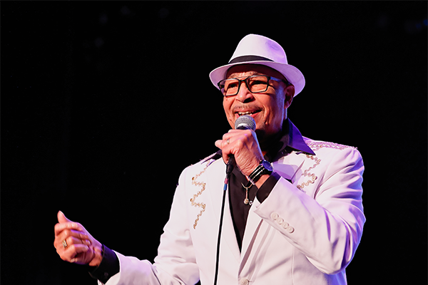 A man in a black shirt with a white suit jacket and fedora singing and snapping into a corded microphone on a stage.