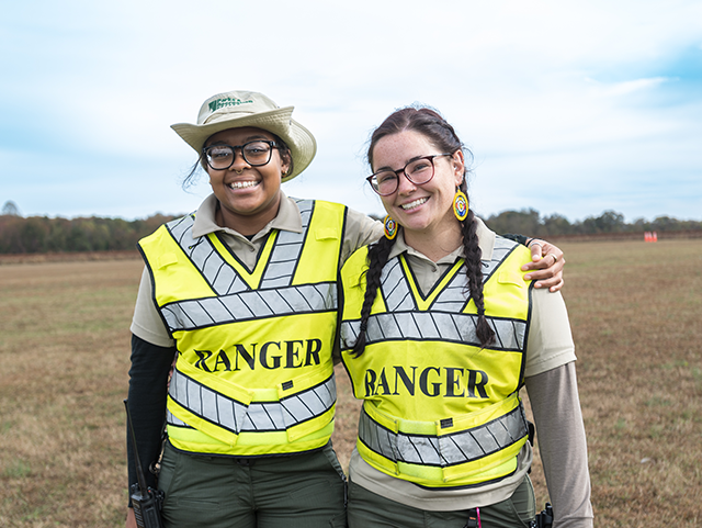 Two women standing next to each other with an arm around each other. They are both smiling and wearing high-visibility vests with the word "Ranger" on the front. They are standing in a field with a far away tree line on the horizon.